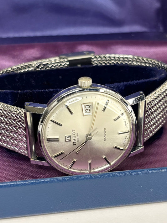 A Tissot Basel watch with a silver-toned stainless steel mesh strap and silver dial, resting inside a blue presentation box with a purple velvet lining.