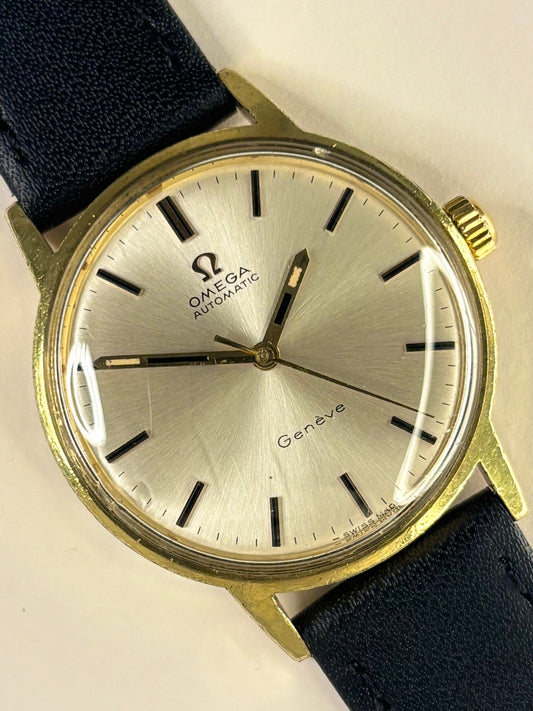 Omega Automatic Geneva watch with a gold-tone case, silver dial, and dark blue leather strap, shown close-up on a light background.