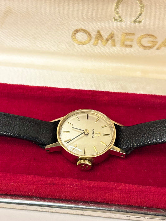 A vintage Omega watch with a gold-toned case, cream dial, and black leather strap, resting on a red velvet cushion inside an Omega-branded presentation box.