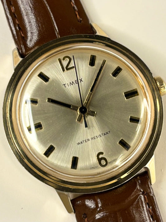 A vintage Timex wristwatch with a round silver dial, black Arabic numerals, black hour markers, and a gold-tone case, paired with a brown leather strap.