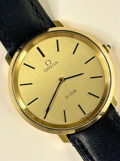 Omega De-Ville wristwatch with a gold dial, gold-tone case, and a black leather strap, shown on a plain light background.