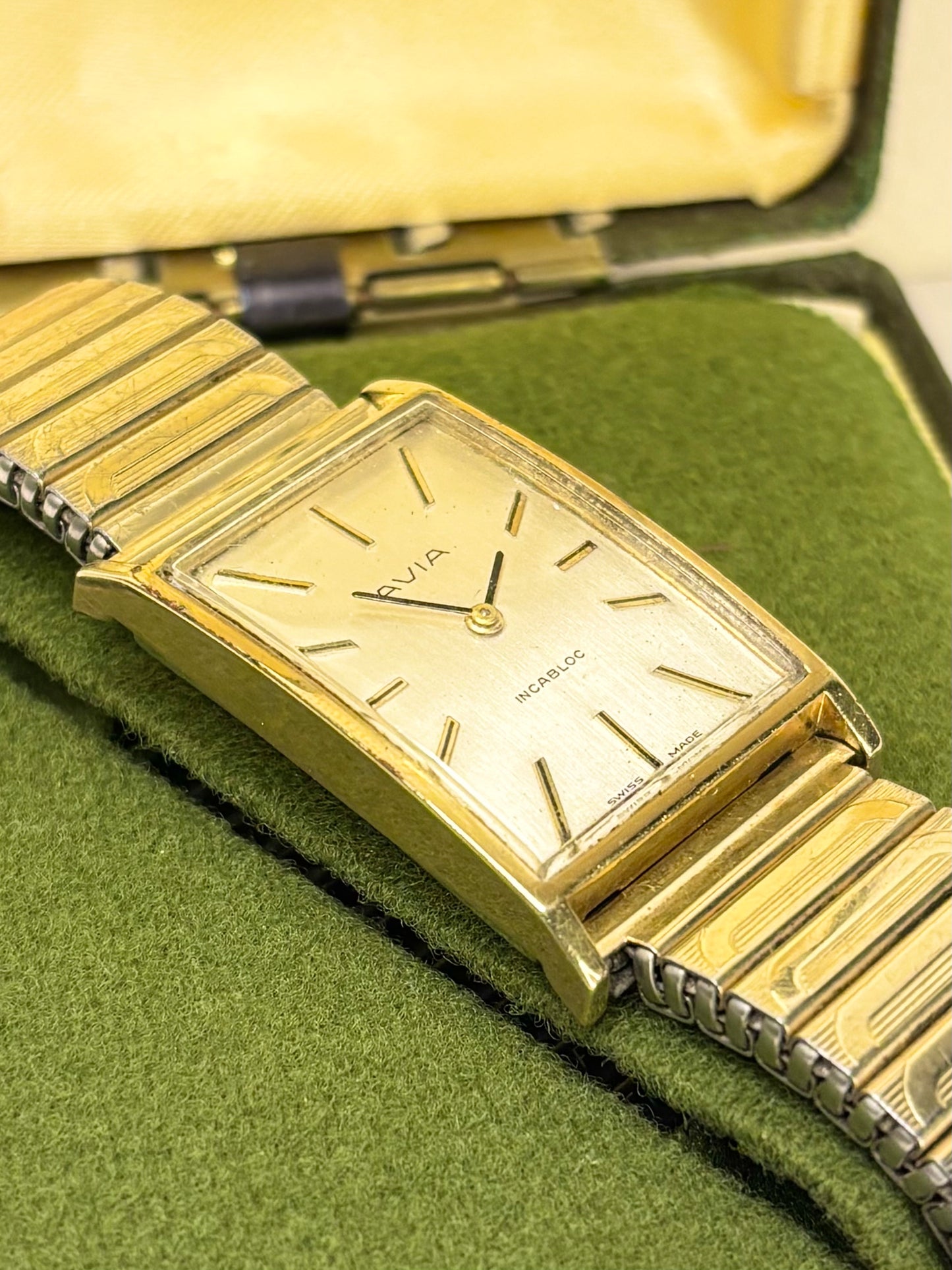 A vintage Omega Swatch wristwatch with a gold-tone rectangular case and link bracelet, resting on a green felt surface inside an open yellow-green presentation box.