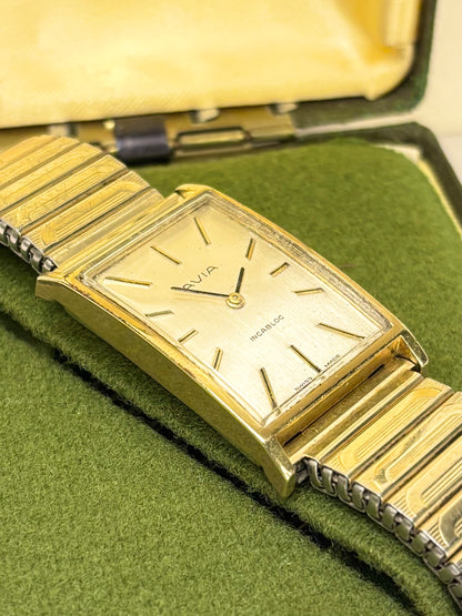 A vintage Omega Swatch wristwatch with a gold-tone rectangular case and link bracelet, resting on a green felt surface inside an open yellow-green presentation box.