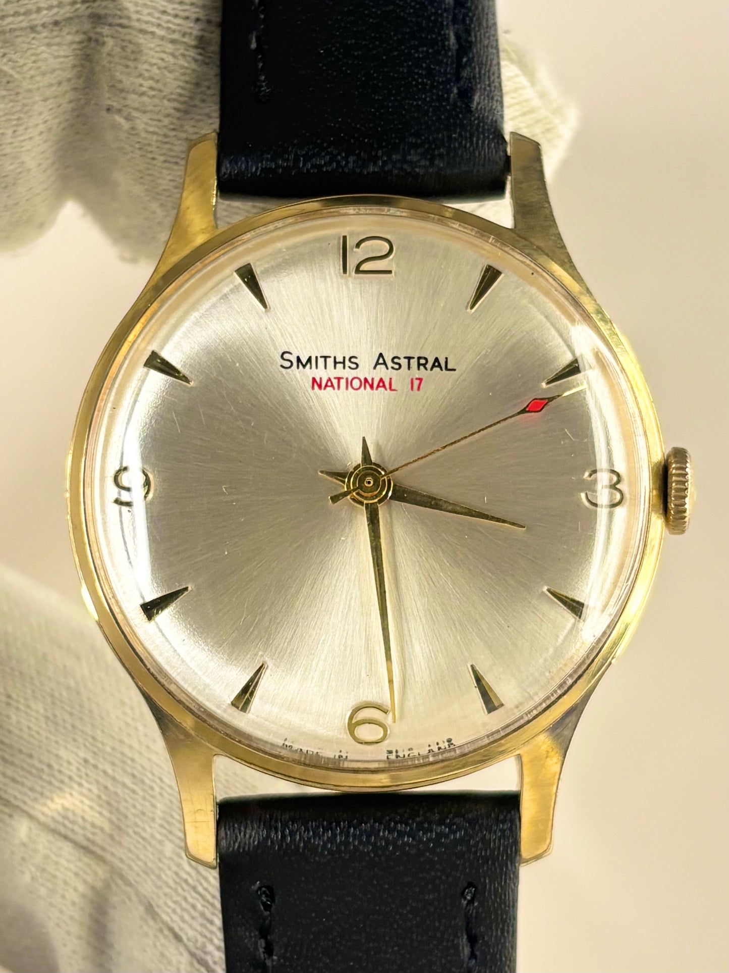 A vintage Smiths Astral National 17 wristwatch with a gold-toned case, silver dial, and black leather strap, held by a hand wearing a white glove.