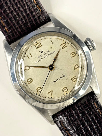 A vintage Rolex Oyster Speeding Precision watch with a silver-toned metal case, a cream-colored dial with gold-tone Arabic numerals, and a dark brown textured leather strap.