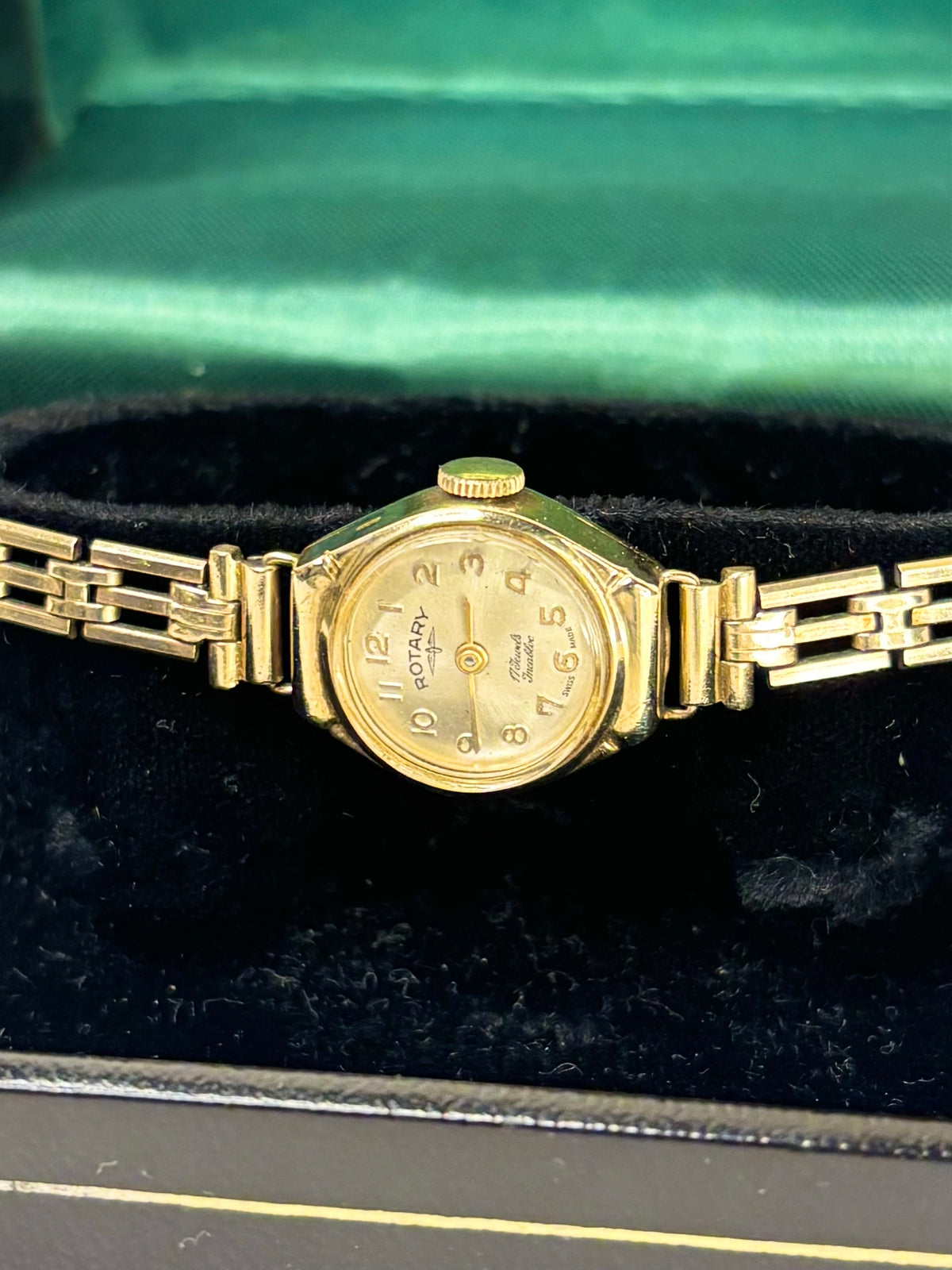 A vintage gold-tone wristwatch with a matching metal link bracelet, resting on a black velvet surface inside a green satin-lined box.