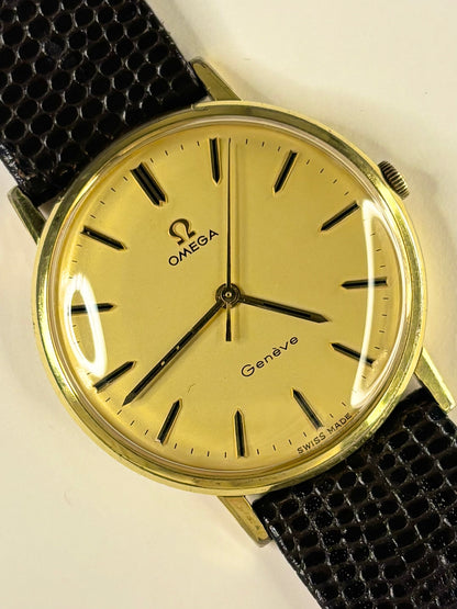 Omega Geneva analog watch with a gold-toned dial, gold case, and a textured black leather strap, shown on a plain light background.