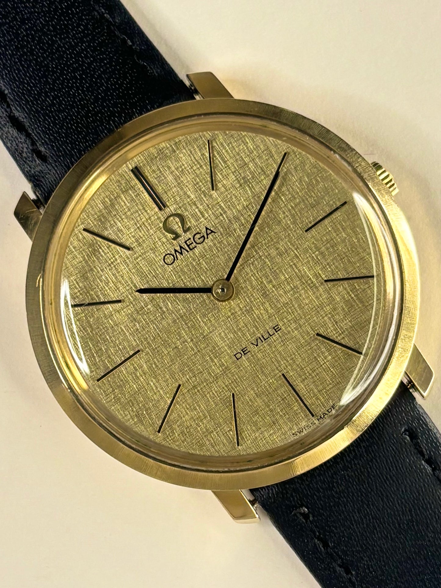 Omega De Ville wristwatch with a gold-toned textured dial, gold case, and a dark navy blue leather strap, shown on a light beige background.