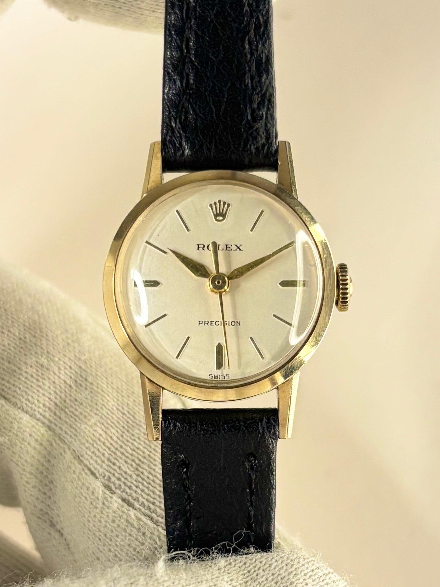 A vintage Rolex Precision watch with a gold case, white dial, and black leather strap, held by a white textured glove against a light beige background.