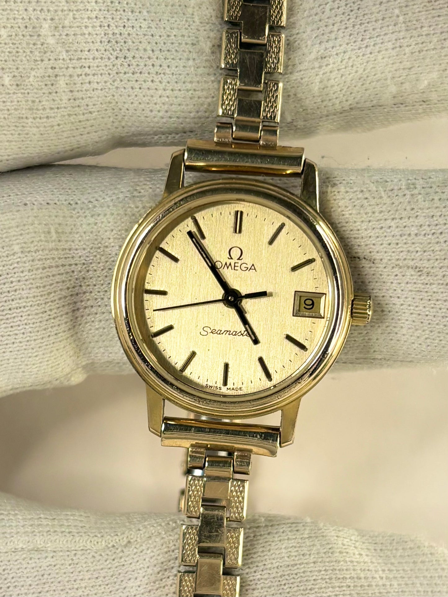 Omega Seamaster stainless steel watch with a cream-colored dial and a matching metal link bracelet, displayed on a textured white fabric background.