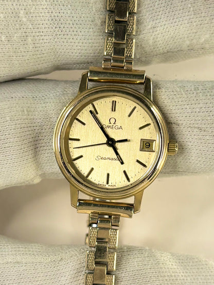 Omega Seamaster stainless steel watch with a cream-colored dial and a matching metal link bracelet, displayed on a textured white fabric background.