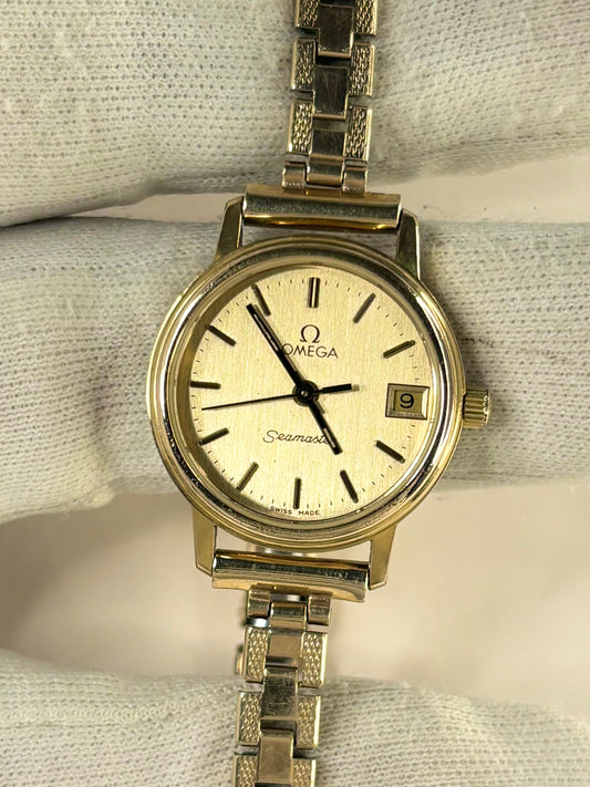 Omega Seamaster stainless steel watch with a cream-colored dial and a matching metal link bracelet, displayed on a textured white fabric background.