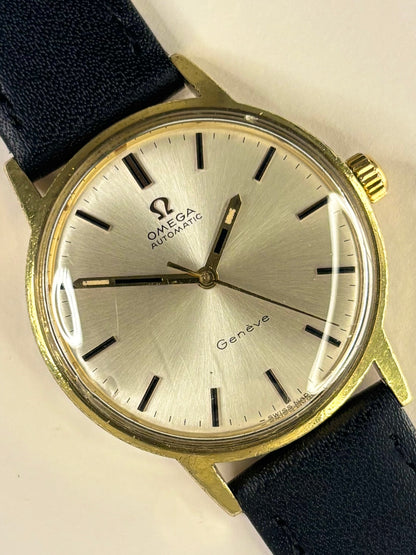 Omega Automatic Geneva watch with a gold-tone case, silver dial, and dark blue leather strap, shown close-up on a light background.