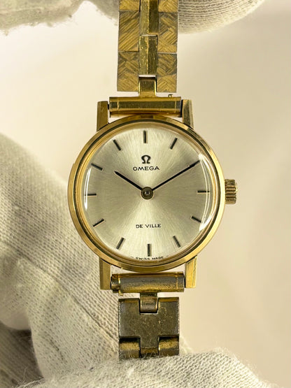 An Omega De Ville wristwatch with a gold-tone case and a textured gold-tone link bracelet, resting on a white textured fabric surface.