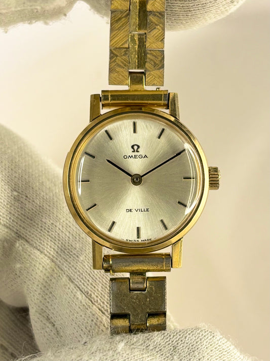 An Omega De Ville wristwatch with a gold-tone case and a textured gold-tone link bracelet, resting on a white textured fabric surface.