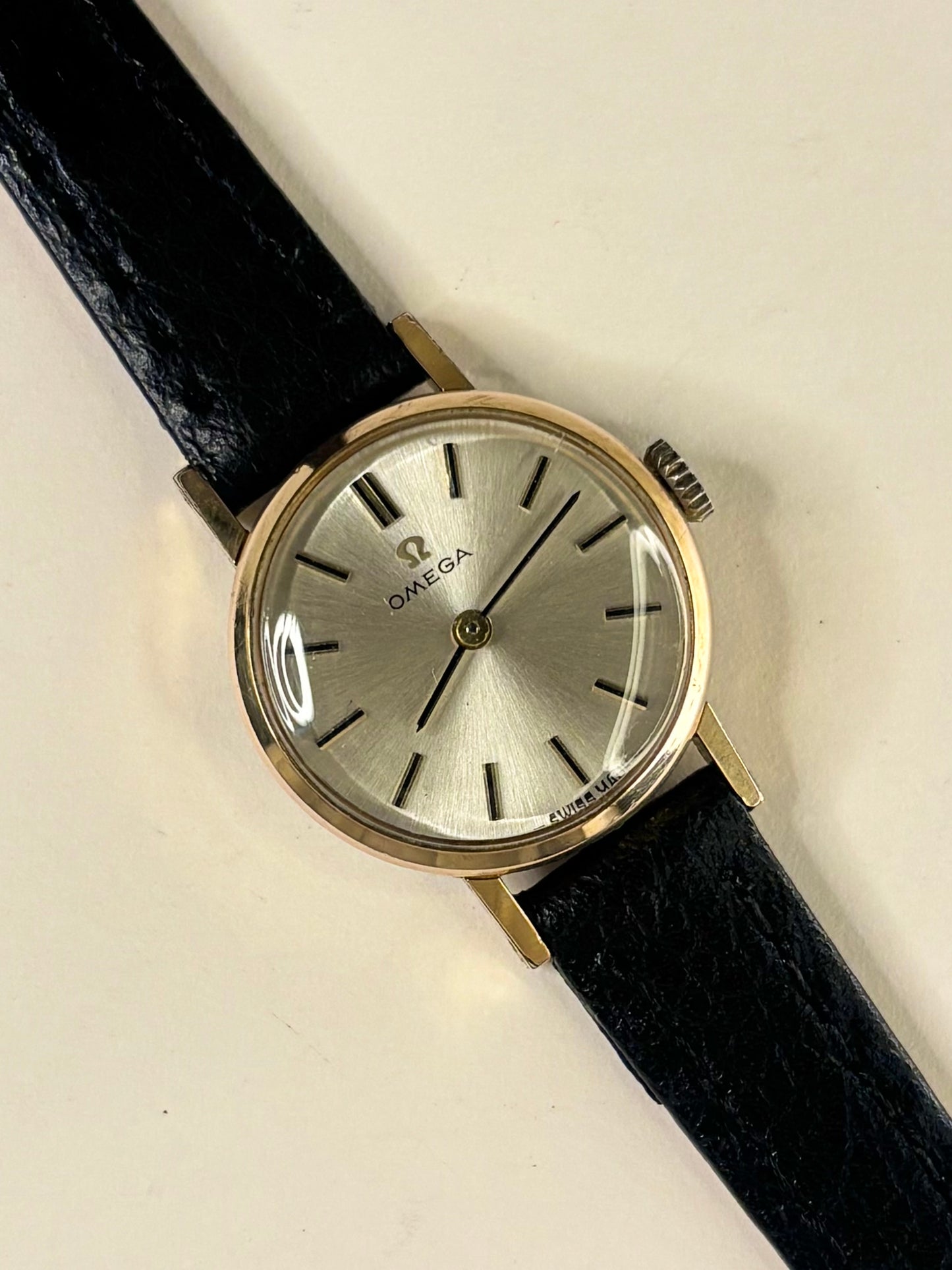 An Omega wristwatch with a round silver dial, gold-tone case, and a black leather strap, shown on a plain off-white background.