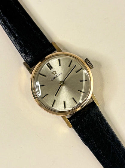 An Omega wristwatch with a round silver dial, gold-tone case, and a black leather strap, shown on a plain off-white background.