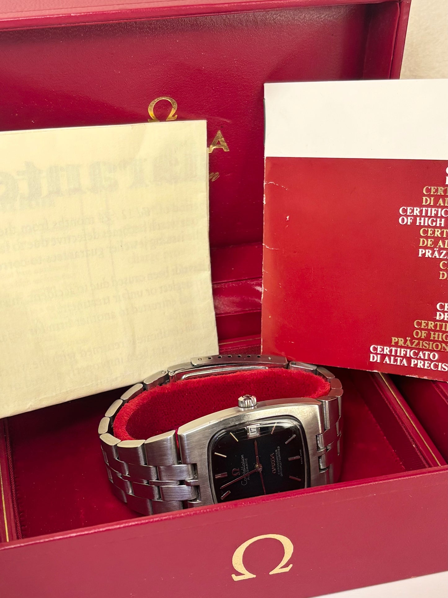 An Omega wristwatch with a stainless steel link bracelet and a black dial, displayed inside its red presentation box with certificates and papers.