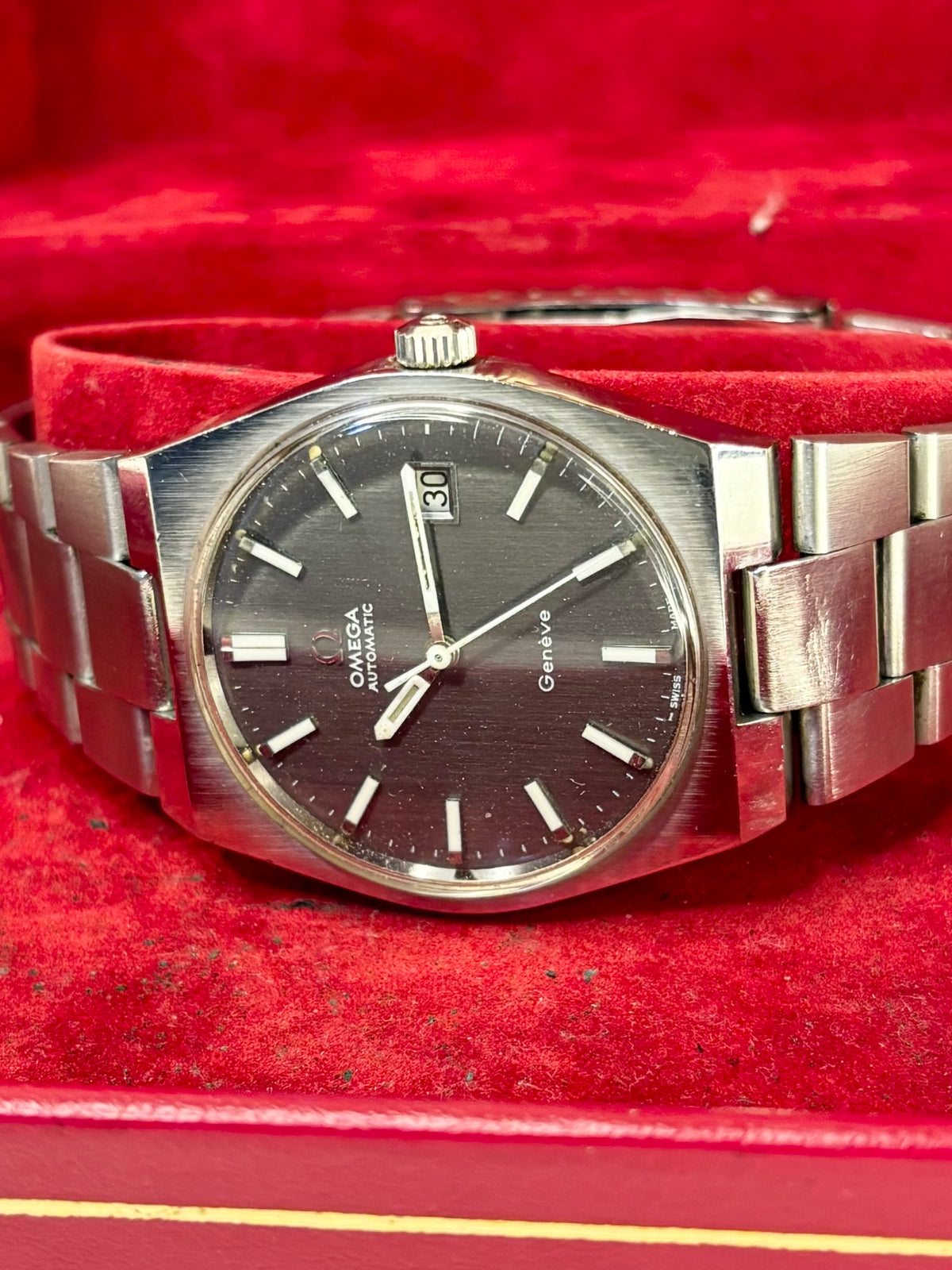 A vintage Omega x Swatch Genedive wristwatch with a brushed silver-tone metal bracelet and a dark dial, displayed inside a red velvet-lined box.