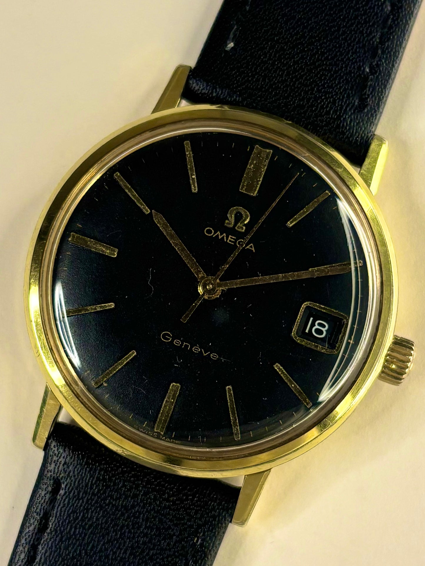 Omega Geneva wristwatch with a gold-tone case, black dial, and black leather strap, shown close-up against a light beige background.