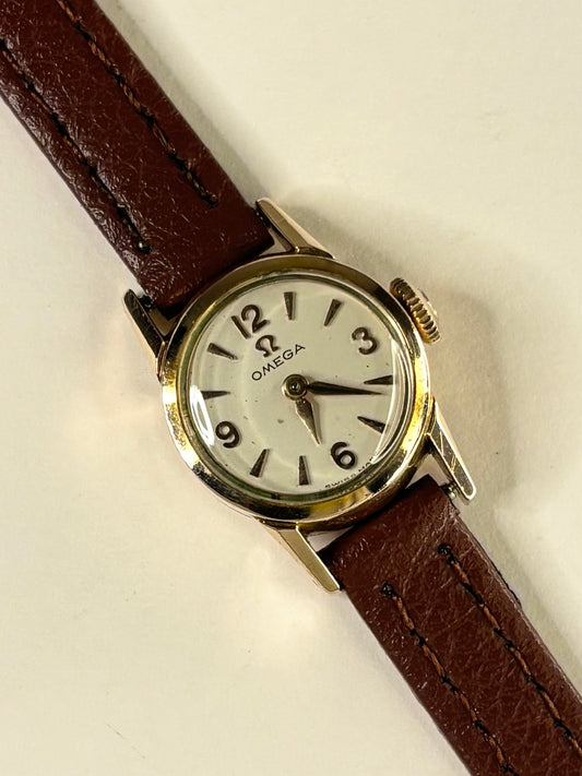 Vintage Omega women's watch with a white dial, gold-tone case, and brown leather strap, shown diagonally on a light beige background.