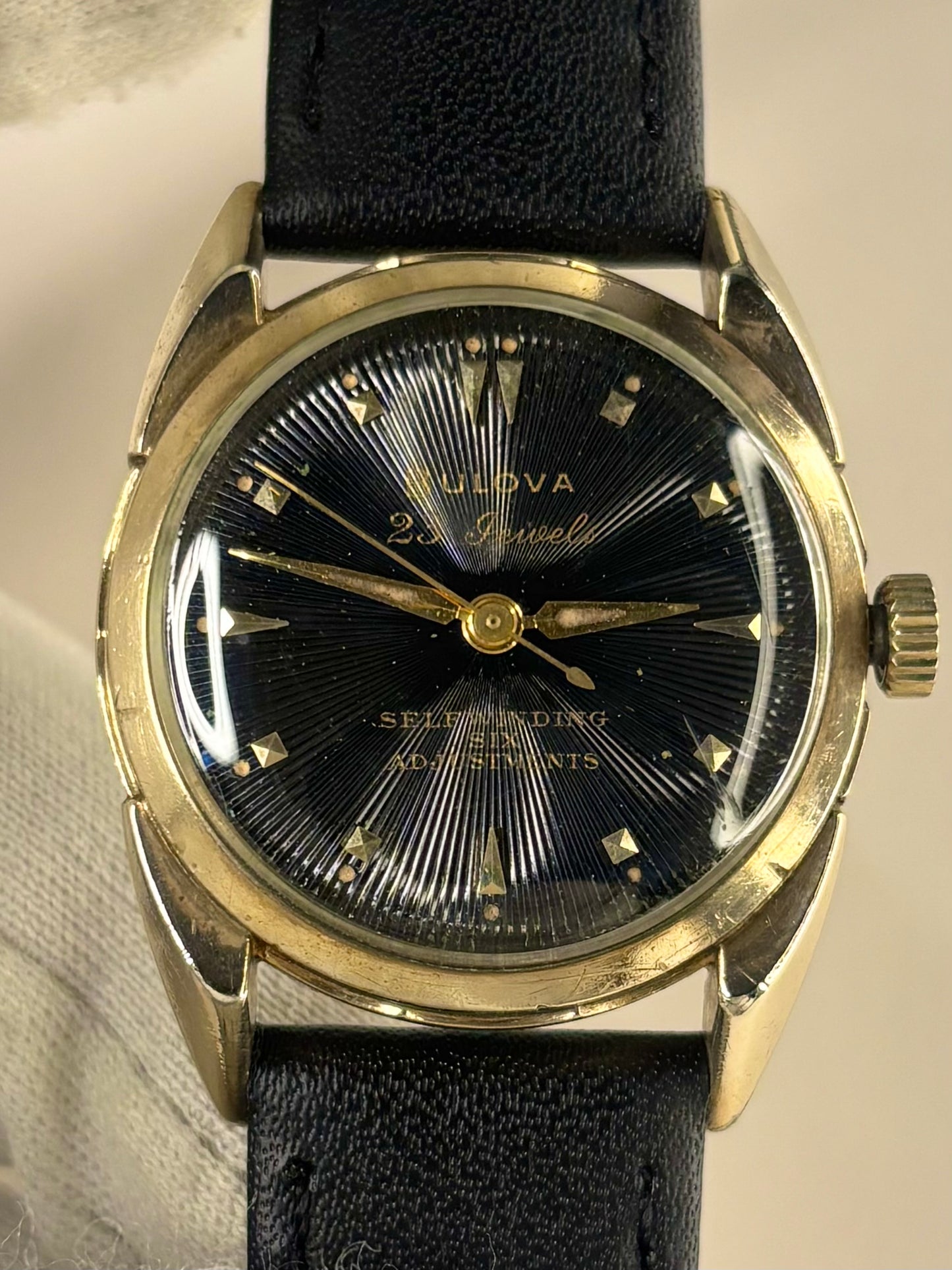 A vintage Bulova 22 Jewels Self-Winding 60m watch with a black sunburst dial, gold-tone case, and black leather strap.