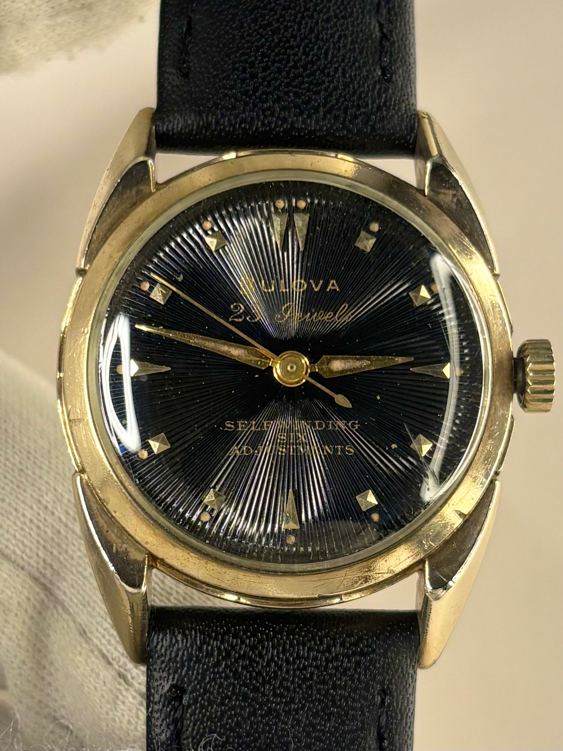 A vintage Bulova 22 Jewels Self-Winding 60m watch with a black sunburst dial, gold-tone case, and black leather strap.