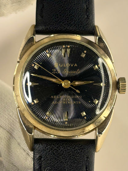 A vintage Bulova 22 Jewels Self-Winding 60m watch with a black sunburst dial, gold-tone case, and black leather strap.