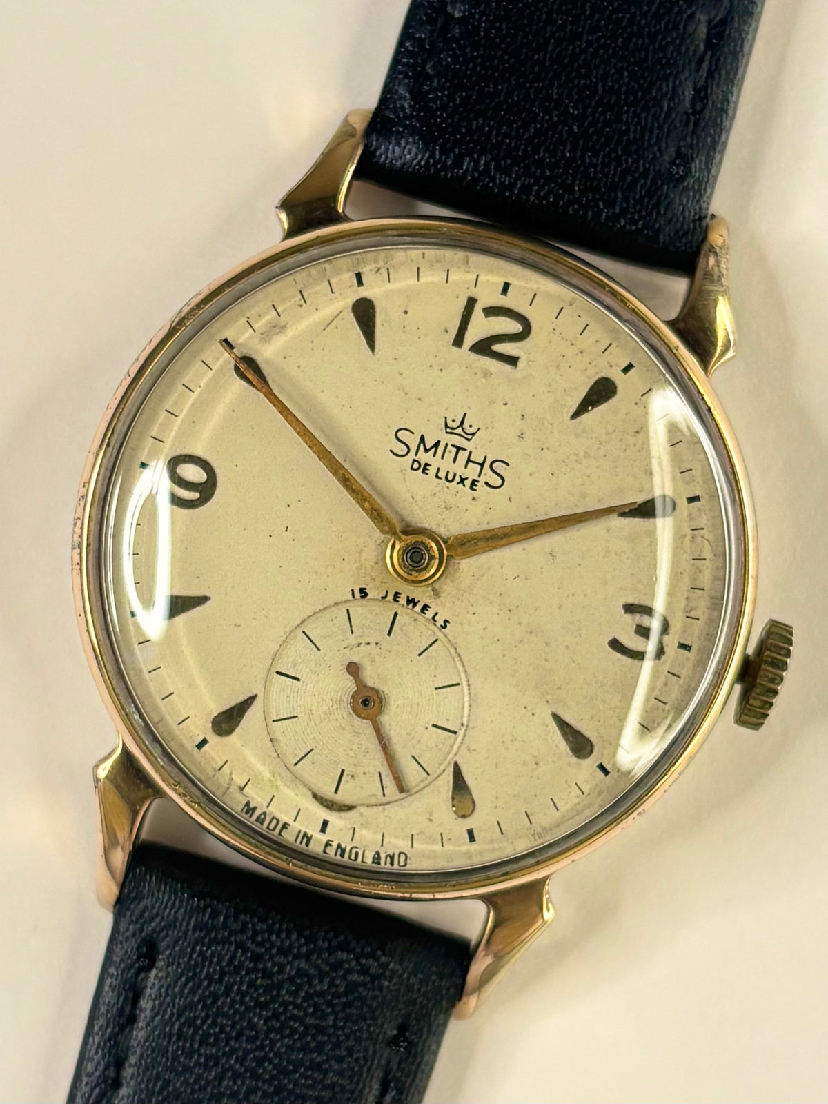A vintage Smiths Colombe wristwatch with a round gold-tone case, cream-colored dial, and a dark blue leather strap, shown against a plain light background.