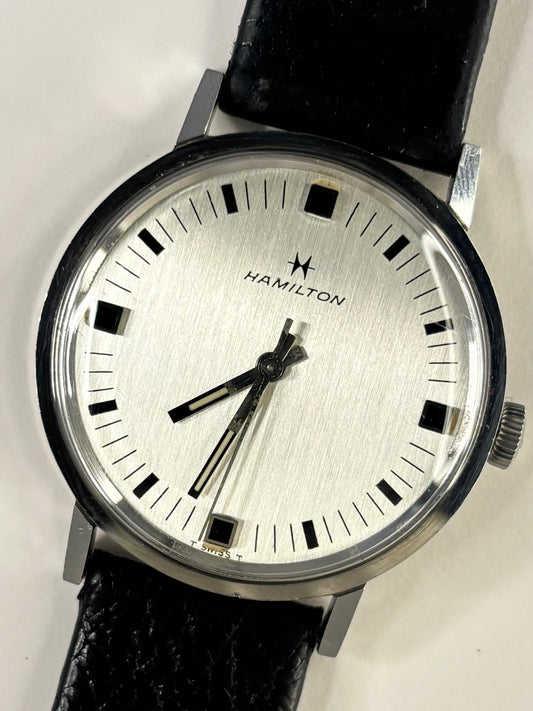 Hamilton wristwatch with a silver dial, black markers and hands, and a black leather strap, shown close-up against a light background.